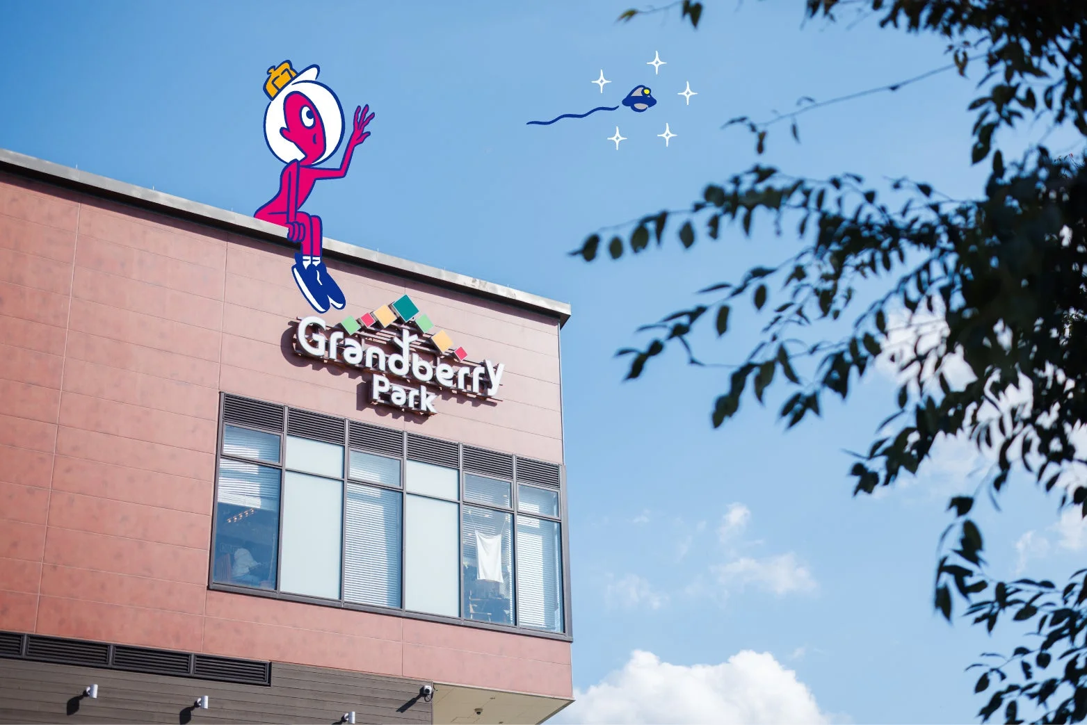 青空の下、「Grandberry Park」の建物に座るピンク色のキャラクターが手を振っており、その上空には光る小さな物体が飛んでいる様子。右側には木の葉が写り込んでいます。