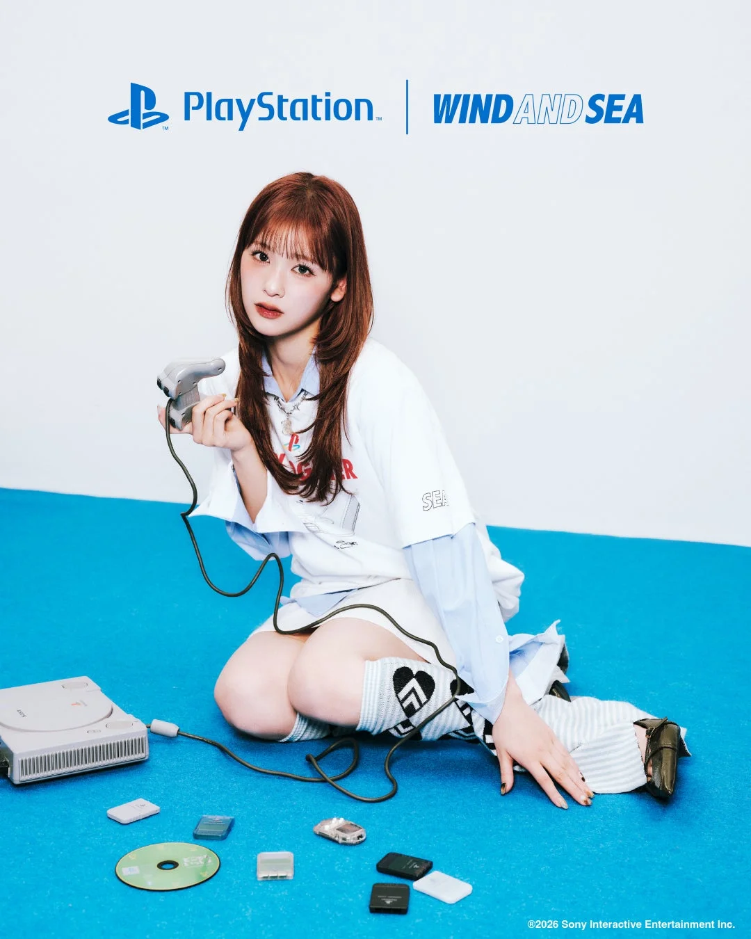 青い床に座った若い女性がPlayStationのコントローラーを手にしています。彼女の周りには初代PlayStation本体やゲーム関連のアイテムが散らばっており、上部にはPlayStationとWIND AND SEAのロゴが見えます。両ブランドのコラボレーションを想起させるプロモーション画像です。