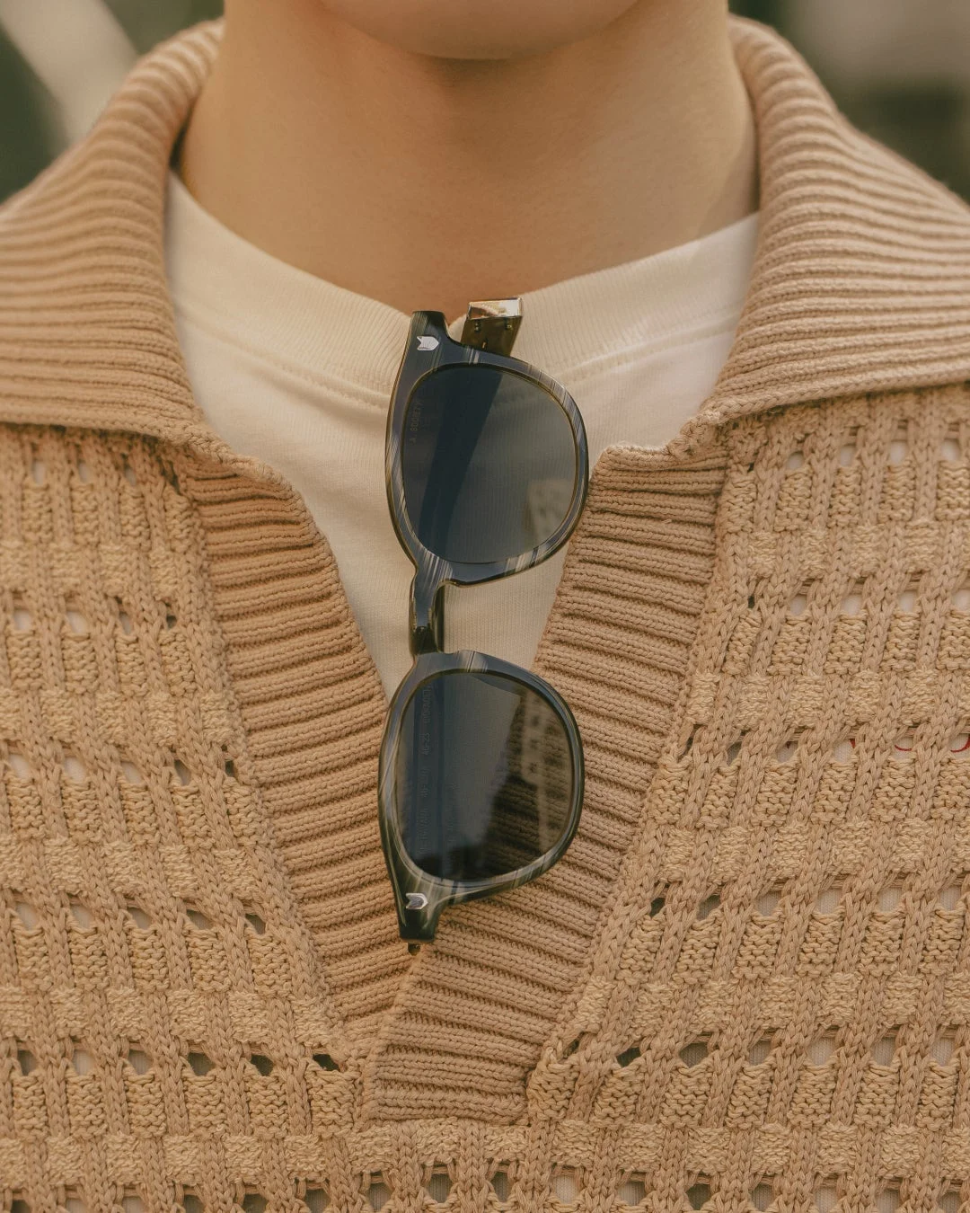 ベージュのVネックニットセーターと白Tシャツを着用した人物の首元のクローズアップ。サングラスが襟元に挟まれている