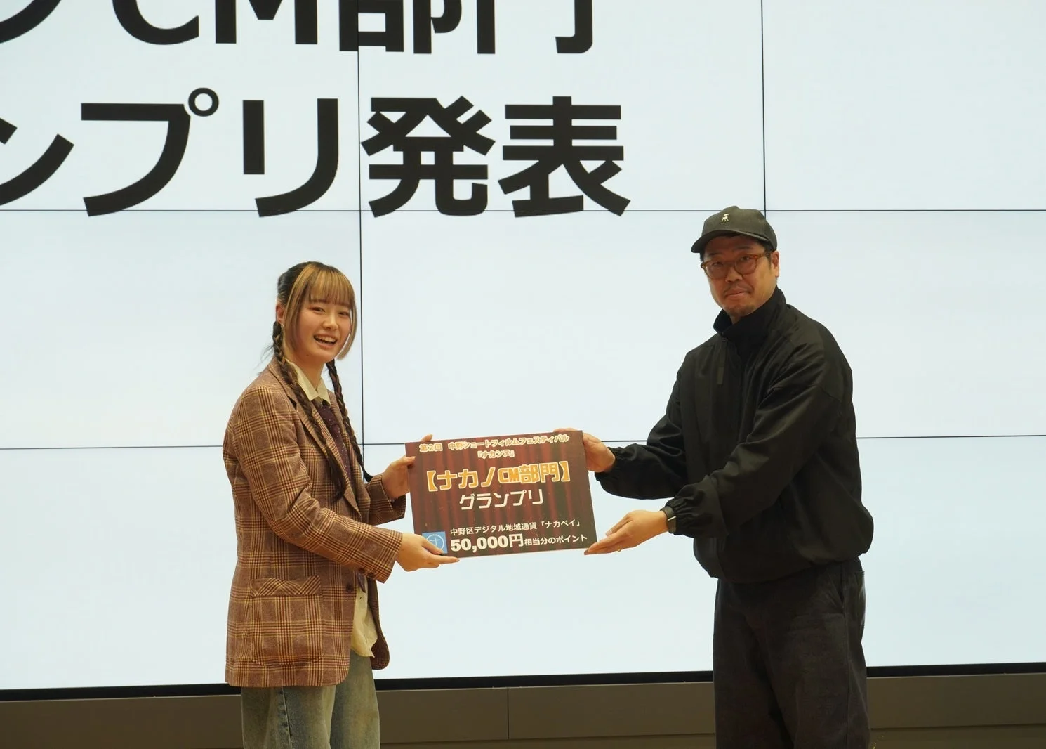 中野ショートフィルムフェスティバル「ナカノア」の授賞式で、一人の女性が男性から「ナカノ/ロ部門」のグランプリ賞状を受け取っています。賞品は中野区デジタル地域通貨「ナカペイ」50,000円相当分のポイントで、背景のスクリーンには「CM部門グランプリ発表」と表示されています。