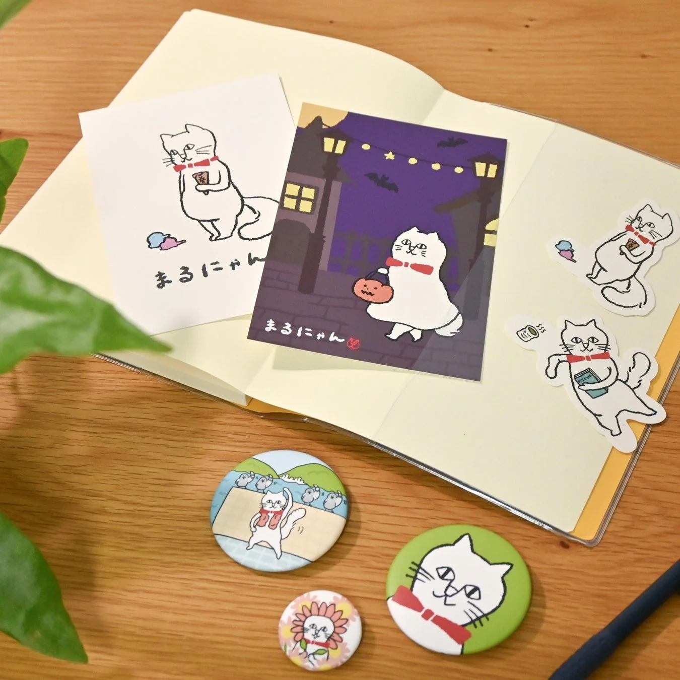 白い猫のキャラクター「まるにゃん」が描かれたポストカード、ステッカー、缶バッジなどの文房具が木製のテーブルに置かれています。ハロウィンをテーマにしたデザインも含まれており、全体的に可愛らしい雰囲気です。