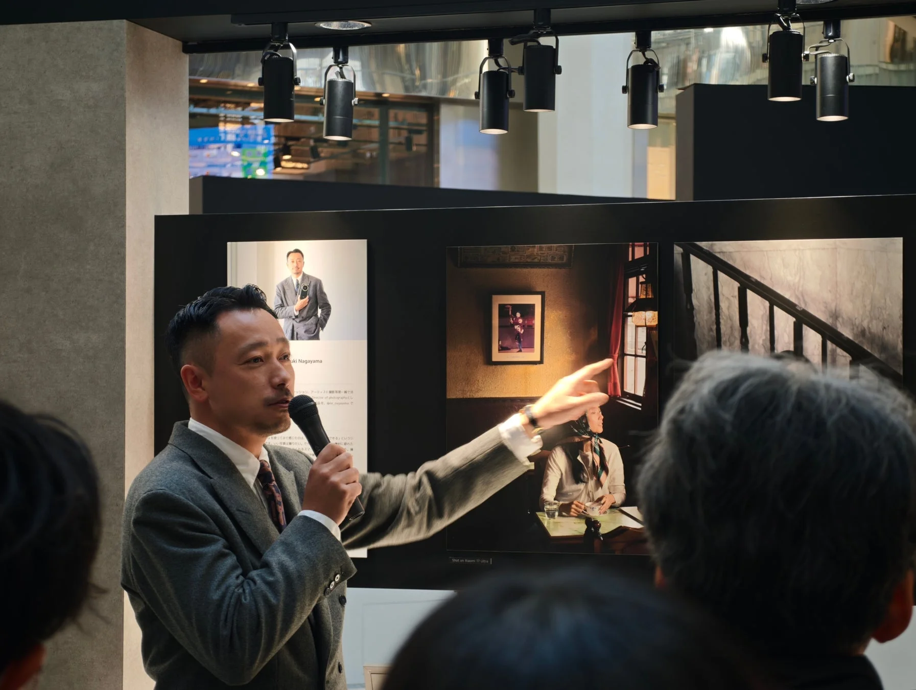 写真展のような会場で、スーツ姿の男性がマイクを手に、展示されている写真について聴衆に説明しています