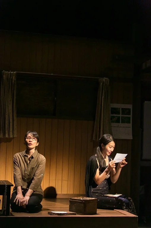 薄暗い舞台で、二人の女性が演技をしている様子。一人は手紙を読み、もう一人は何かを考え込むように見上げています。静かで情緒的な場面です。