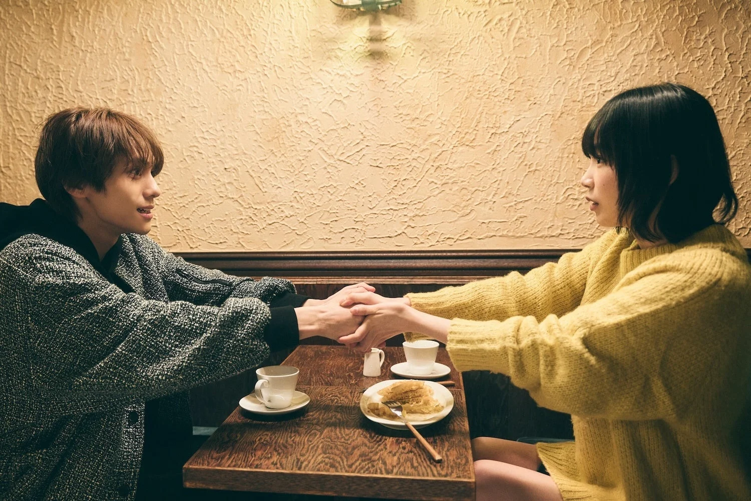 男女がカフェのテーブルで向かい合って座り、手を取り合っている様子。親密な雰囲気で会話を楽しんでいるようだ。テーブルにはコーヒーカップとデザートがある。