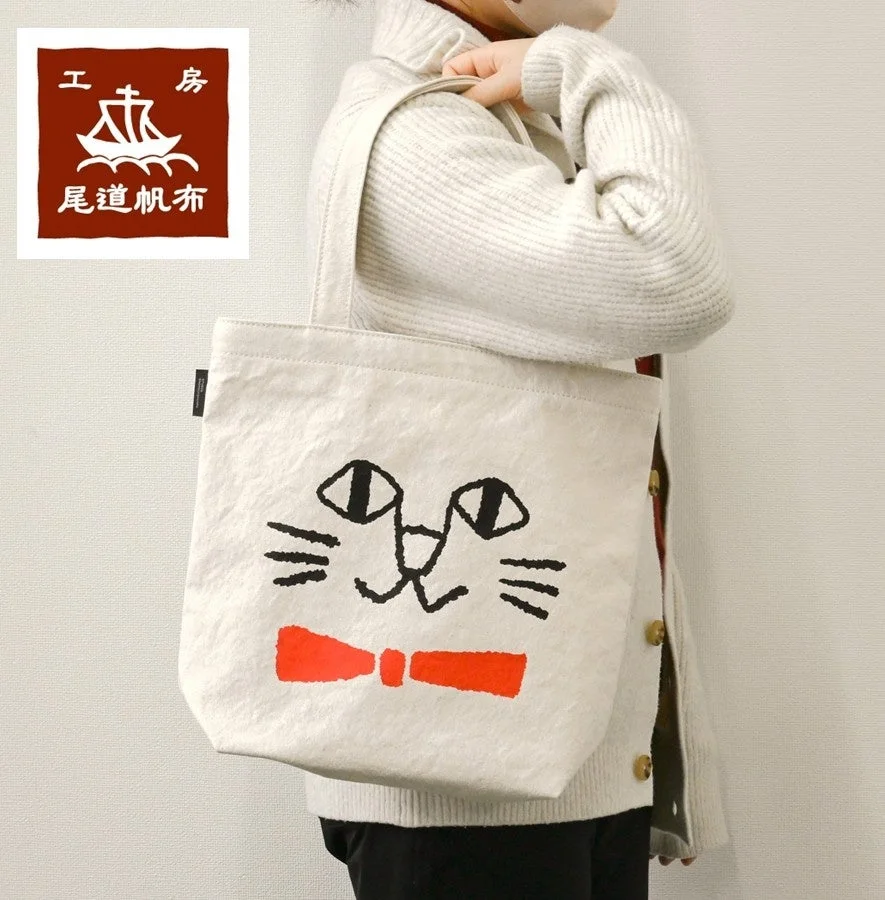 白いニットを着た人物が、猫の顔と赤い蝶ネクタイが描かれた白いトートバッグを持っている画像です。バッグには「尾道帆布」のロゴが見えます。