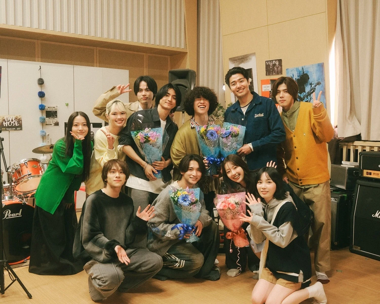音楽スタジオのような場所で、男女11人の若者が笑顔で集合写真を撮っている。多くの人が花束を手に持ち、お祝いムードが漂う記念写真である。