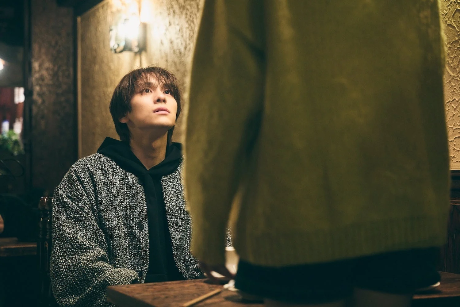 薄暗い室内で、若い男性がテーブルに座り、目の前の人物を真剣な表情で見上げています。黒いパーカーとツイード風のジャケットを着ており、落ち着いた雰囲気の中で会話をしているかのような情景です。