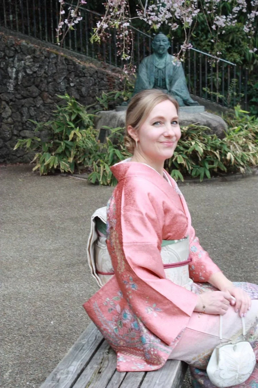 桜が咲く日本の庭園で、ピンクの着物を着た女性がベンチに座り、笑顔でカメラを見つめています。背景には像が見えます。