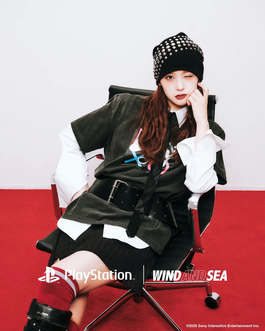 スタッズ付きビーニーと個性的なファッションに身を包んだ若い女性が椅子に座り、カメラに向かってウィンクしている画像です。PlayStationとWIND AND SEAのロゴがあり、コラボレーションを示唆しています。