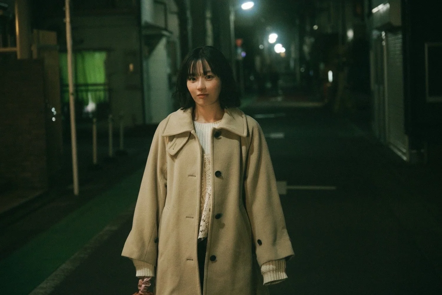 夜の薄暗い街角に立つ若い女性のポートレート。ベージュのコートを着用し、カメラをまっすぐ見つめています。街灯の光が背景にぼんやりと見え、どこか物憂げな雰囲気を醸し出しています。