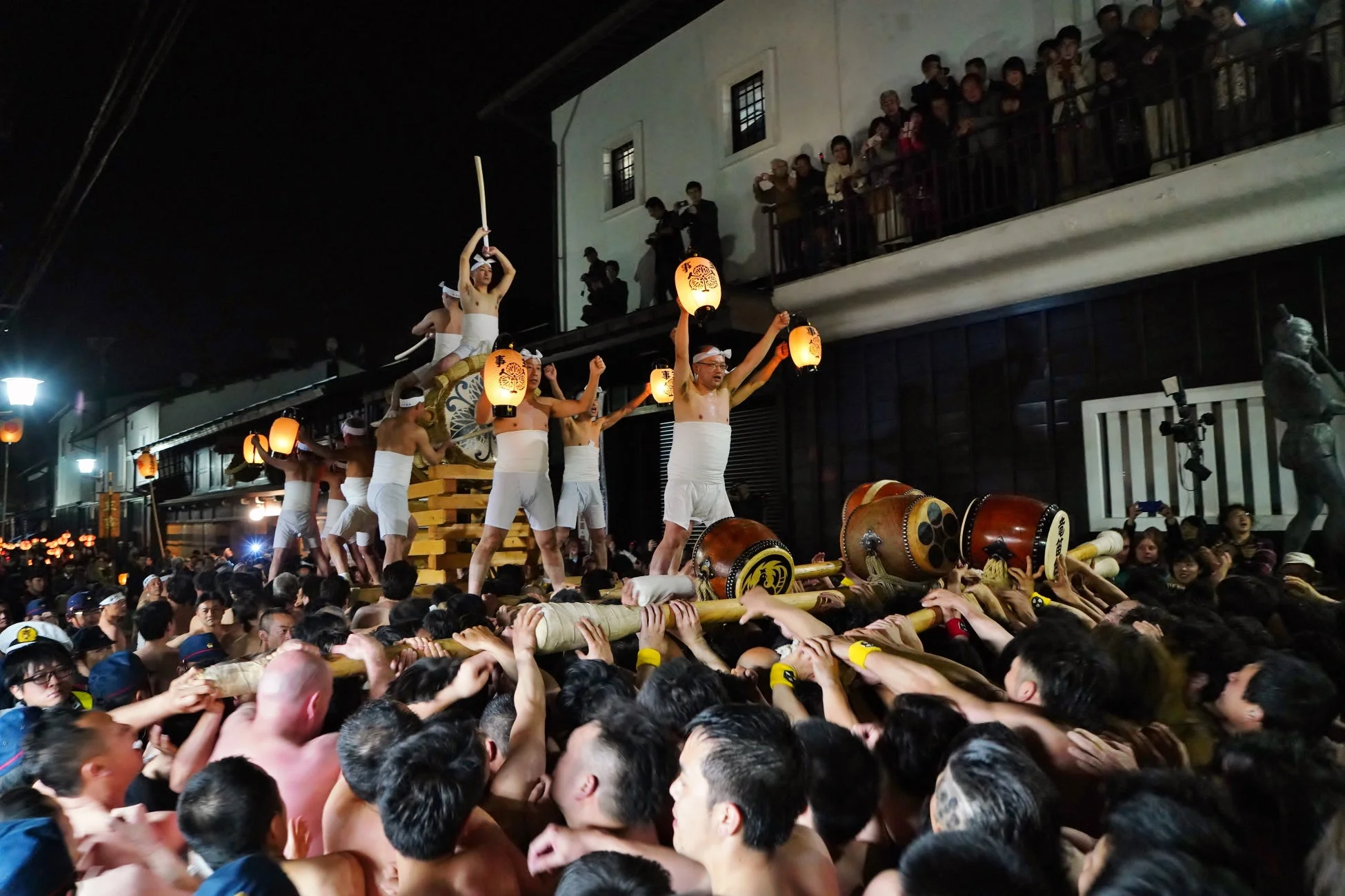 夜の祭り風景。大勢の人々が大きな山車を担ぎ、その上では白装束の男たちが提灯や棒を掲げている。建物のバルコニーからも観客が見守り、活気に満ちた伝統的な祭りの様子がうかがえる。