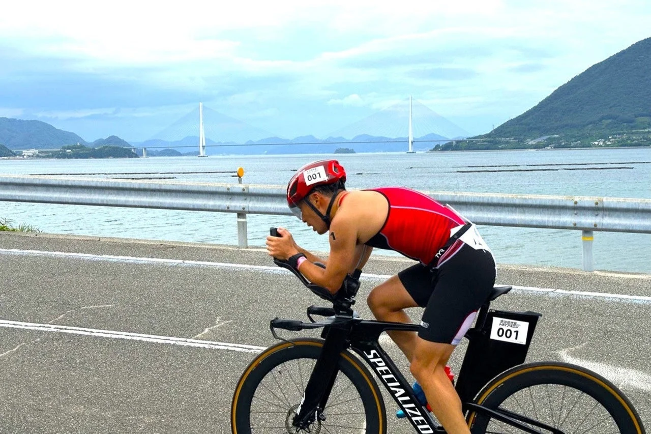 赤いウェアとヘルメットを着用したアスリートが、海沿いの道をタイムトライアルバイクで走行している