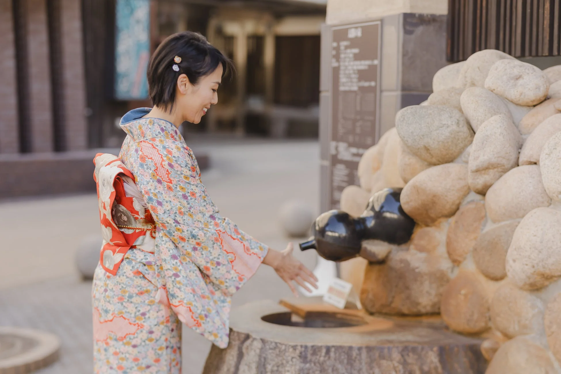 華やかな着物を着た女性が、石造りの足湯のような水場に手を差し伸べています。日本の伝統的な風景の中で、笑顔で穏やかな時間を楽しんでいる様子が伝わります。