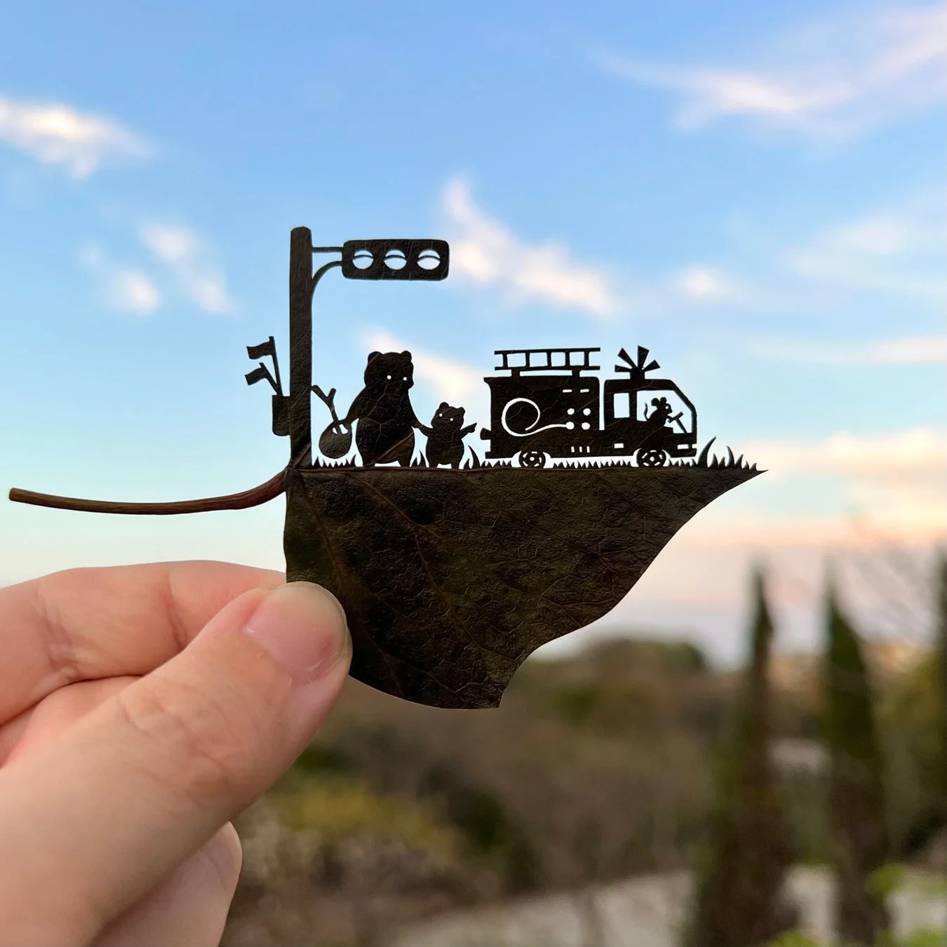 青空を背景に、葉っぱに切り抜かれたクマの親子、消防車、信号機のアート作品が指で持たれています。まるで物語の一場面のような可愛らしいシルエットです。