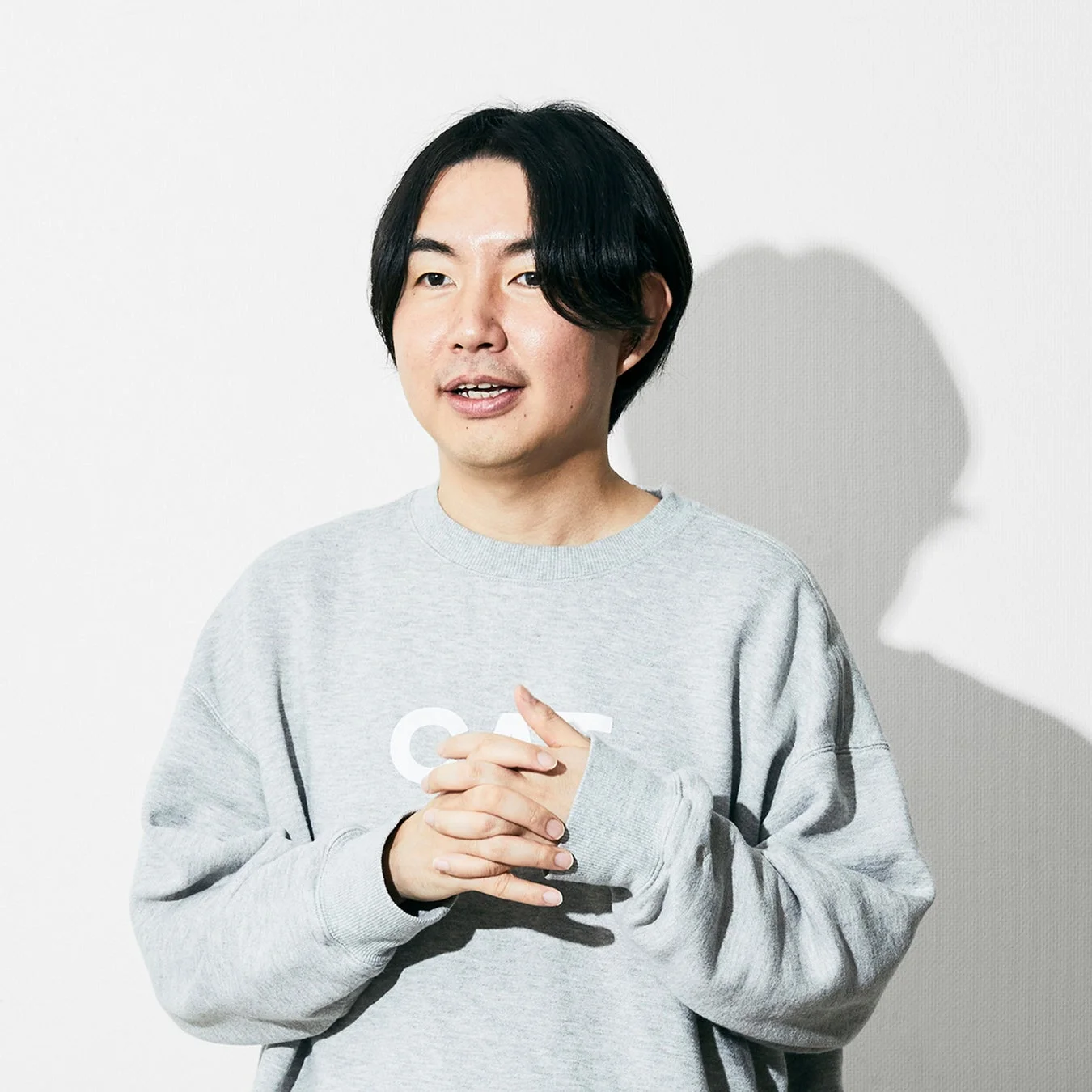 黒髪のアジア人男性がグレーのスウェットシャツを着用し、手を組んでやや左を向いている上半身のポートレート写真です。シンプルな白い背景の前で撮影されています。