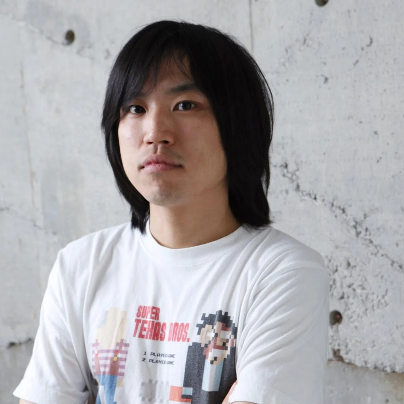 長髪のアジア人男性のポートレート。白いTシャツを着ており、Tシャツには「SUPER TEXAS BROS.」というピクセルアート風のゲームロゴがプリントされています。