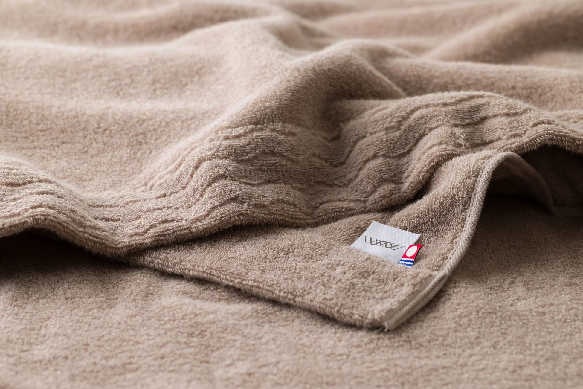 ベージュ色の柔らかそうなタオルが丁寧に折りたたまれています。タオルの表面には独特の織り模様があり、右下には「weavve」と書かれたブランドタグが見えます。暖かく心地よい質感が伝わってきます。
