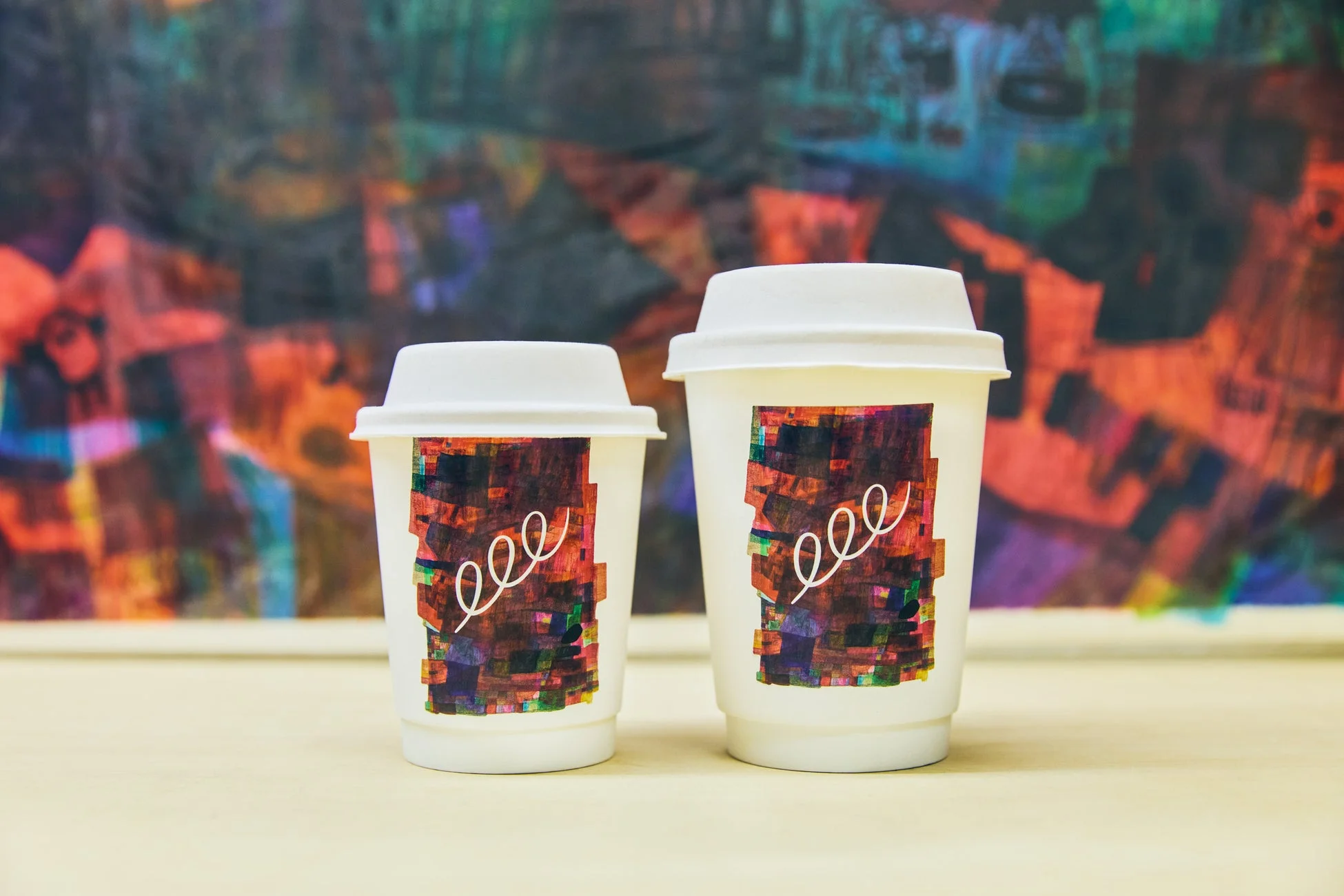 カラフルな抽象画を背景に、デザインラベルが貼られた大小2つの白いテイクアウトカップ