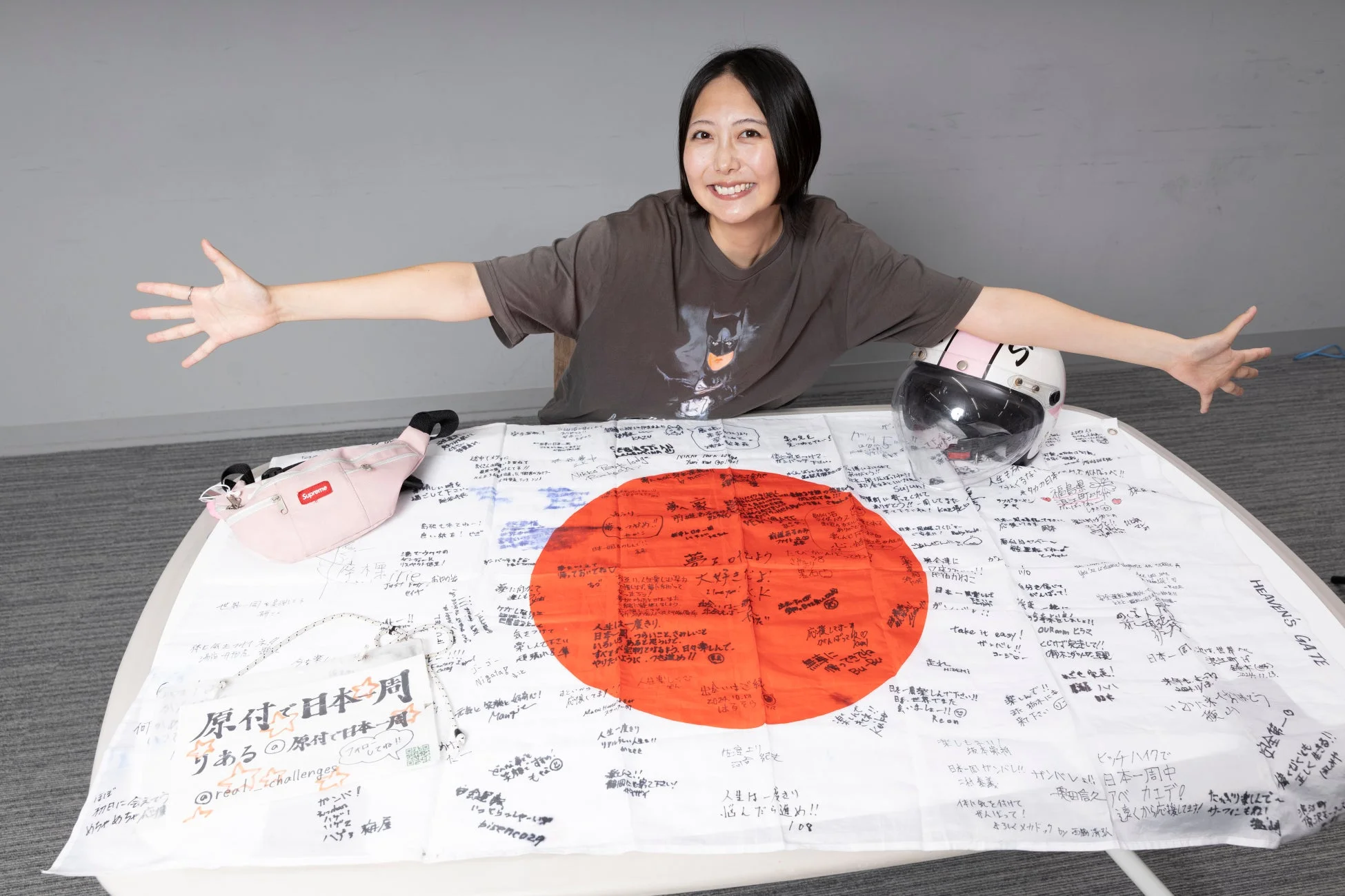 笑顔の女性が、原付での日本一周を応援するメッセージで埋め尽くされた日の丸の旗を広げています。