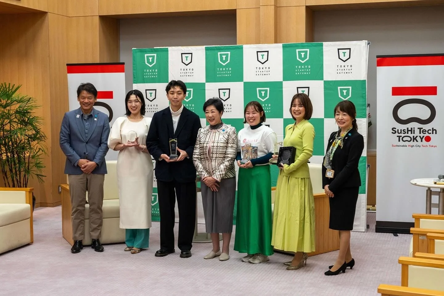 「TOKYO STARTUP GATEWAY」と「SusHi Tech TOKYO」のバナーを背景に、男女7人がトロフィーなどを手に並んでいる集合写真です。