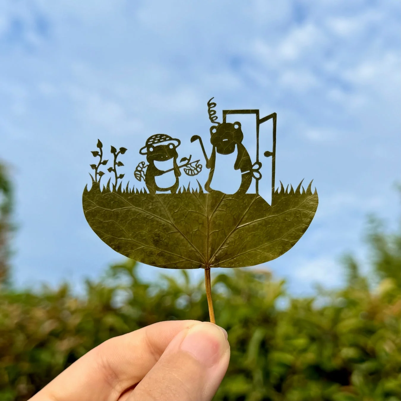 手に持たれた葉っぱに、カエルのキャラクターが二匹描かれた精巧な切り絵アート。一匹は麦わら帽子をかぶり収穫物を持ち、もう一匹はコック帽とエプロン姿で、ドアの前に立つ庭の情景が表現されています。青空と緑を背景にした、創造性豊かな作品です。