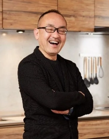 眼鏡をかけたアジア系の男性が、キッチンらしき場所で腕を組み、口を大きく開けて楽しそうに笑っているポートレート写真です。