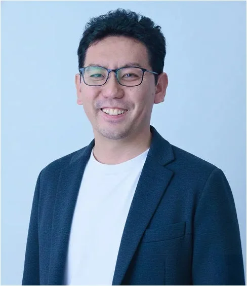 眼鏡をかけたアジア人男性が白いTシャツとダークブルーのジャケットを着て、穏やかな笑顔でカメラを見ている上半身のポートレート