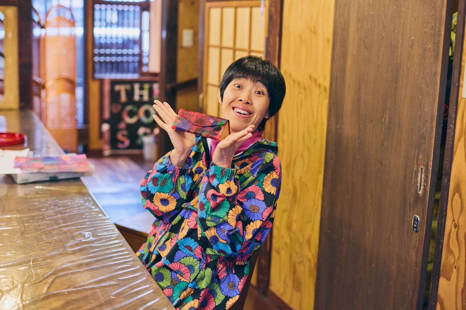 短い黒髪のアジア人女性が、カラフルな花柄のジャケットを着て木製のカウンターに座り、小さな柄物のケースを両手で持ちながら、驚きと喜びの表情を浮かべている様子