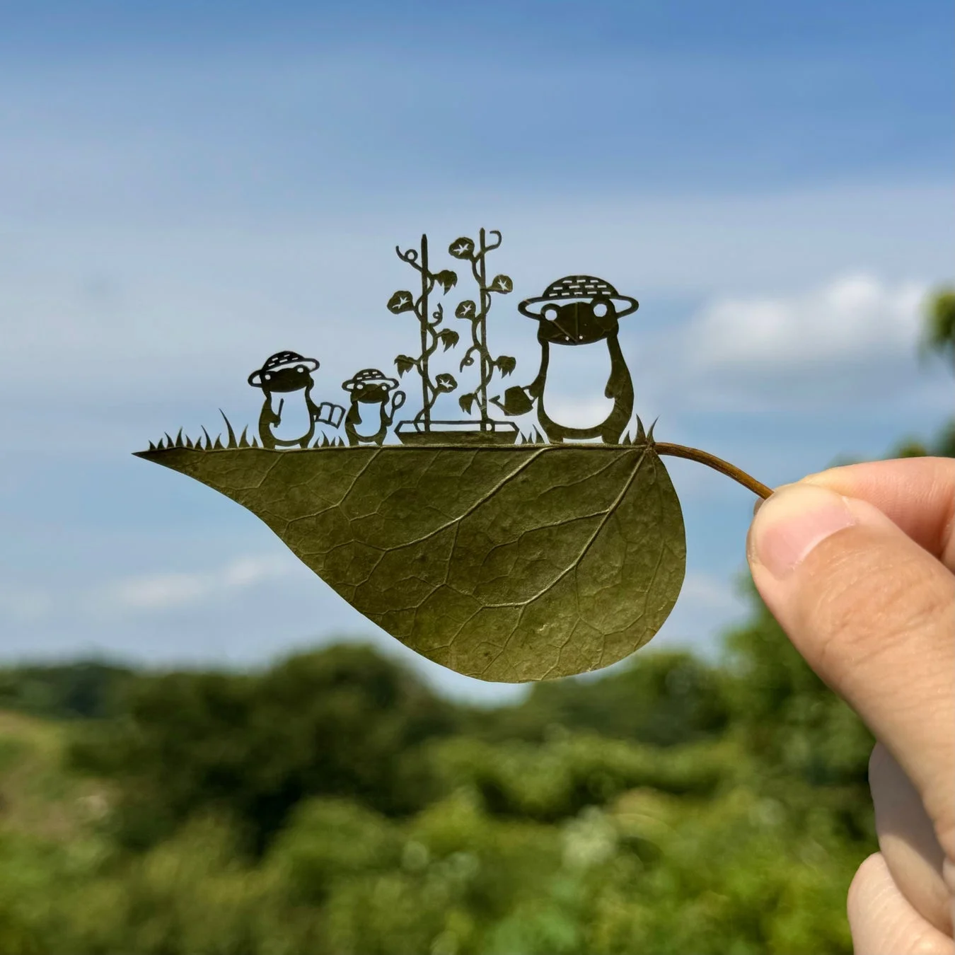 青空を背景に、手に持たれた葉っぱの切り絵。麦わら帽子をかぶったペンギンのようなキャラクターたちが、朝顔のガーデニングを楽しむ様子が精巧に描かれている。自然とアートが融合した、可愛らしく創造的な作品。