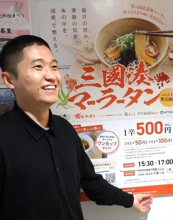 笑顔の男性が「三国湊マーラータン」のポスターの前で宣伝しています。