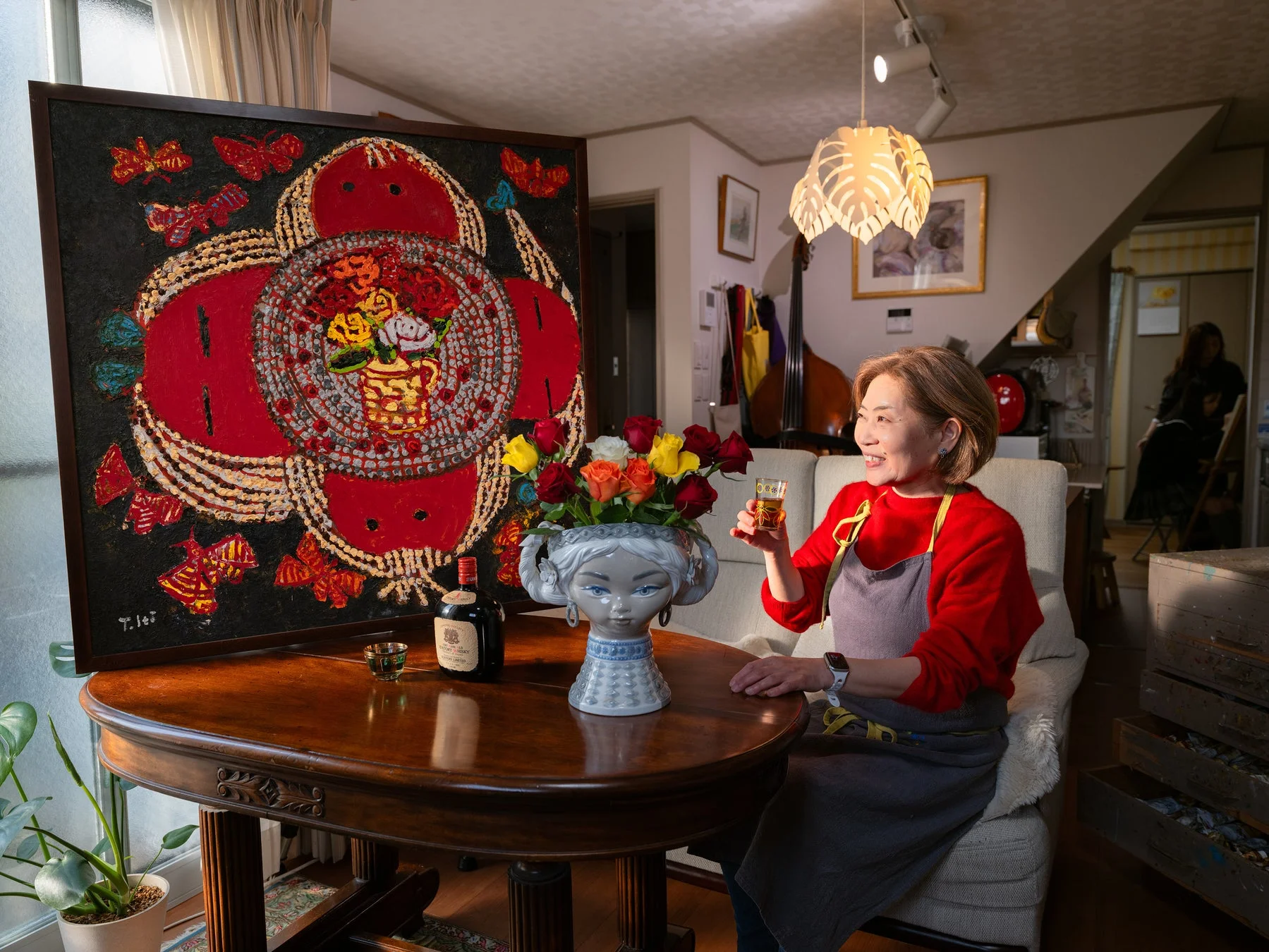 赤いセーターの女性が飲み物を手に絵画の前で微笑む様子