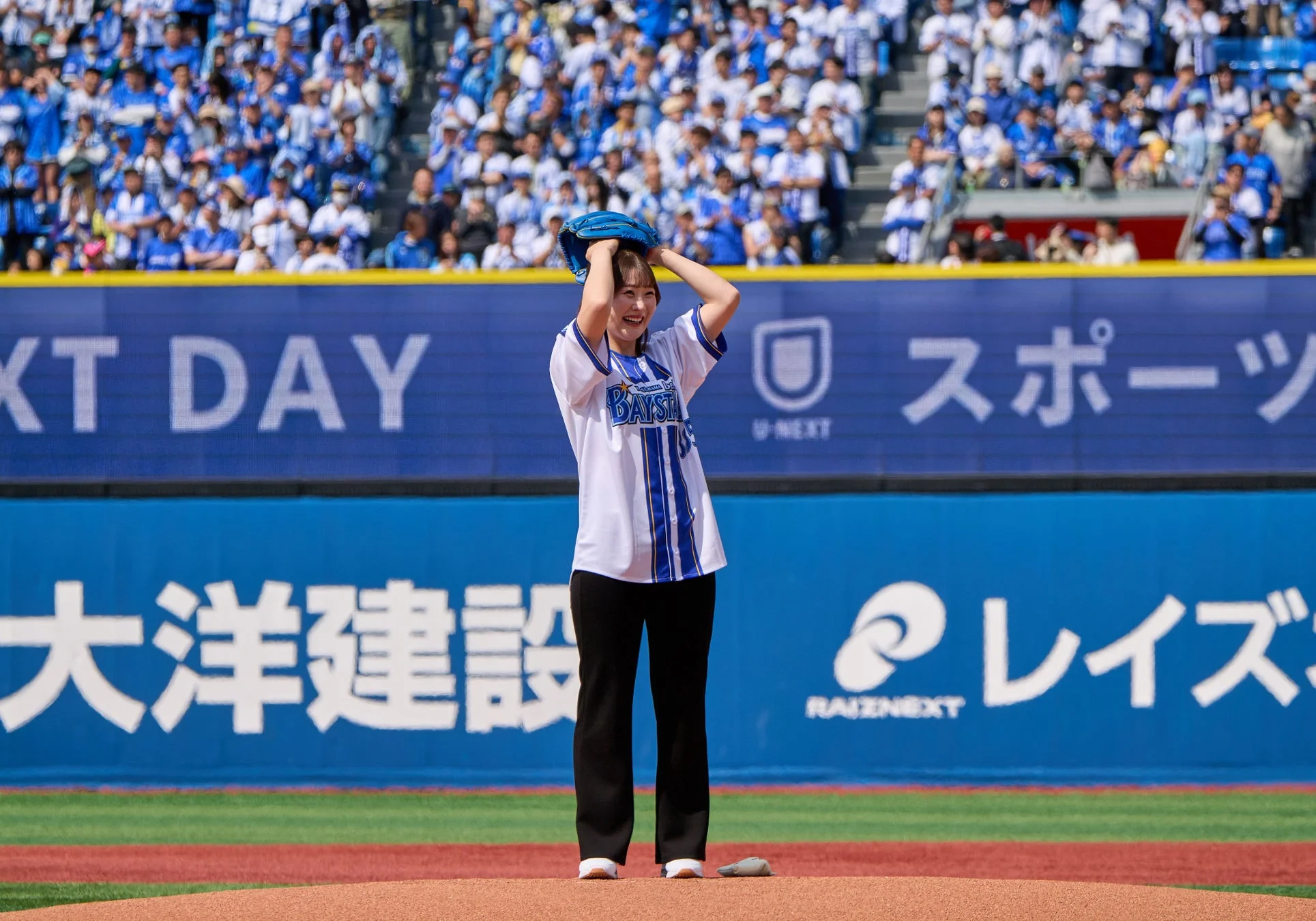 野球場のマウンドに立つ女性が笑顔でポーズをとっています