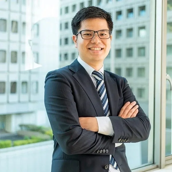 青いストライプのネクタイと眼鏡をかけたビジネスマンが、オフィスらしき窓を背景に腕を組んで笑顔で立っています。