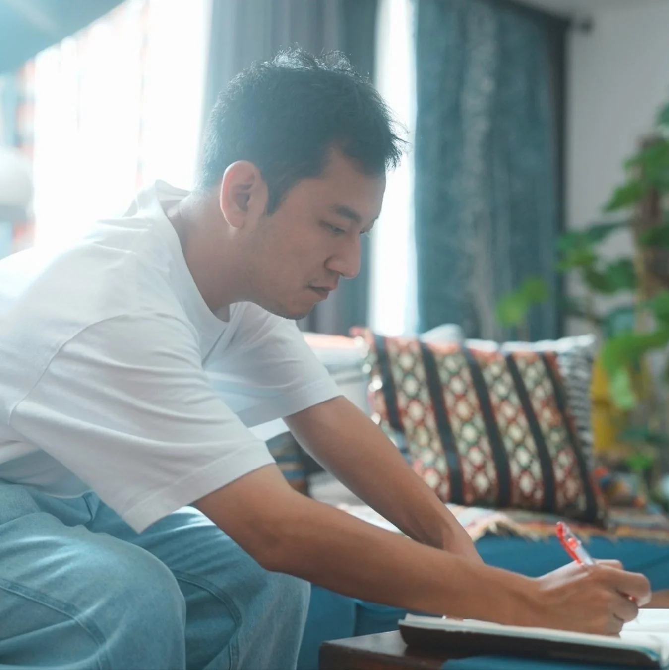 白いTシャツとジーンズを着用した男性が、明るい室内でノートに集中して何かを書き込んでいる様子。