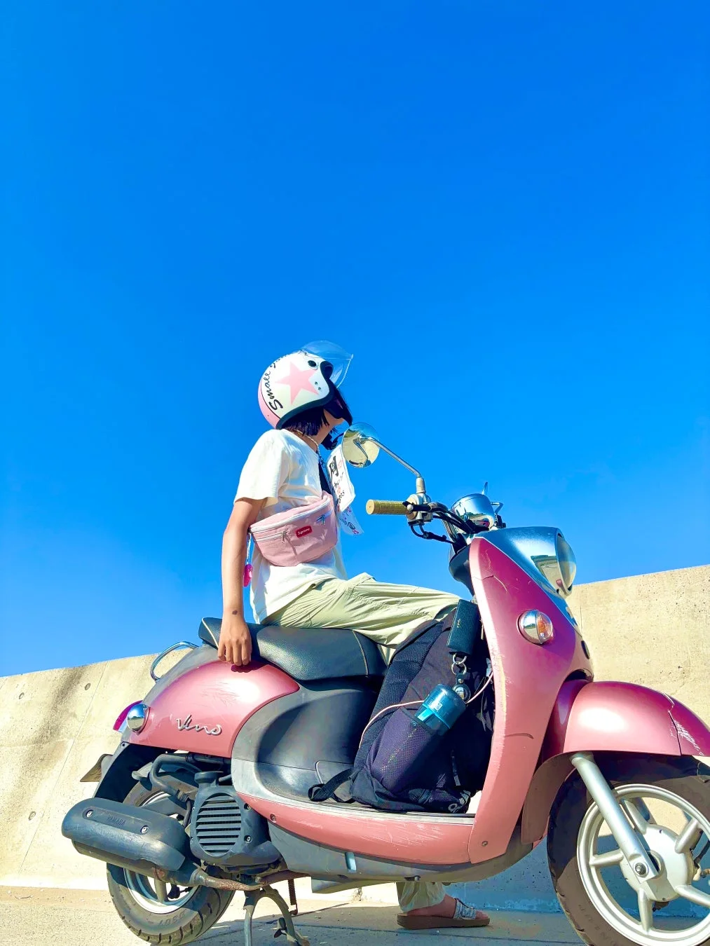 晴れた青空の下、ピンク色のスクーターに座り、星柄のヘルメットをかぶった人物が上を見上げている様子。