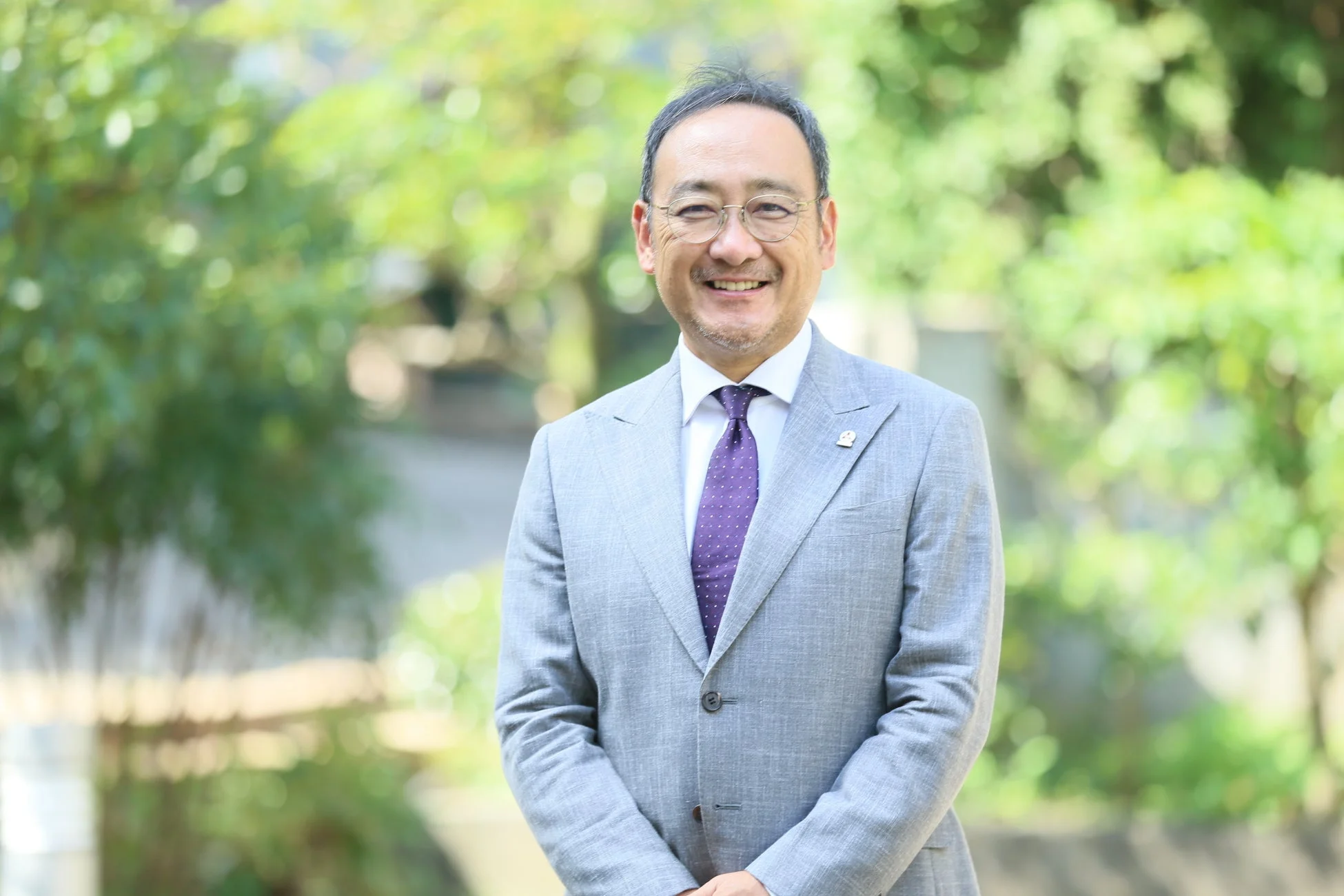 明るい笑顔の男性がライトグレーのスーツと紫のネクタイを着用し、緑豊かな屋外を背景にポーズをとっています