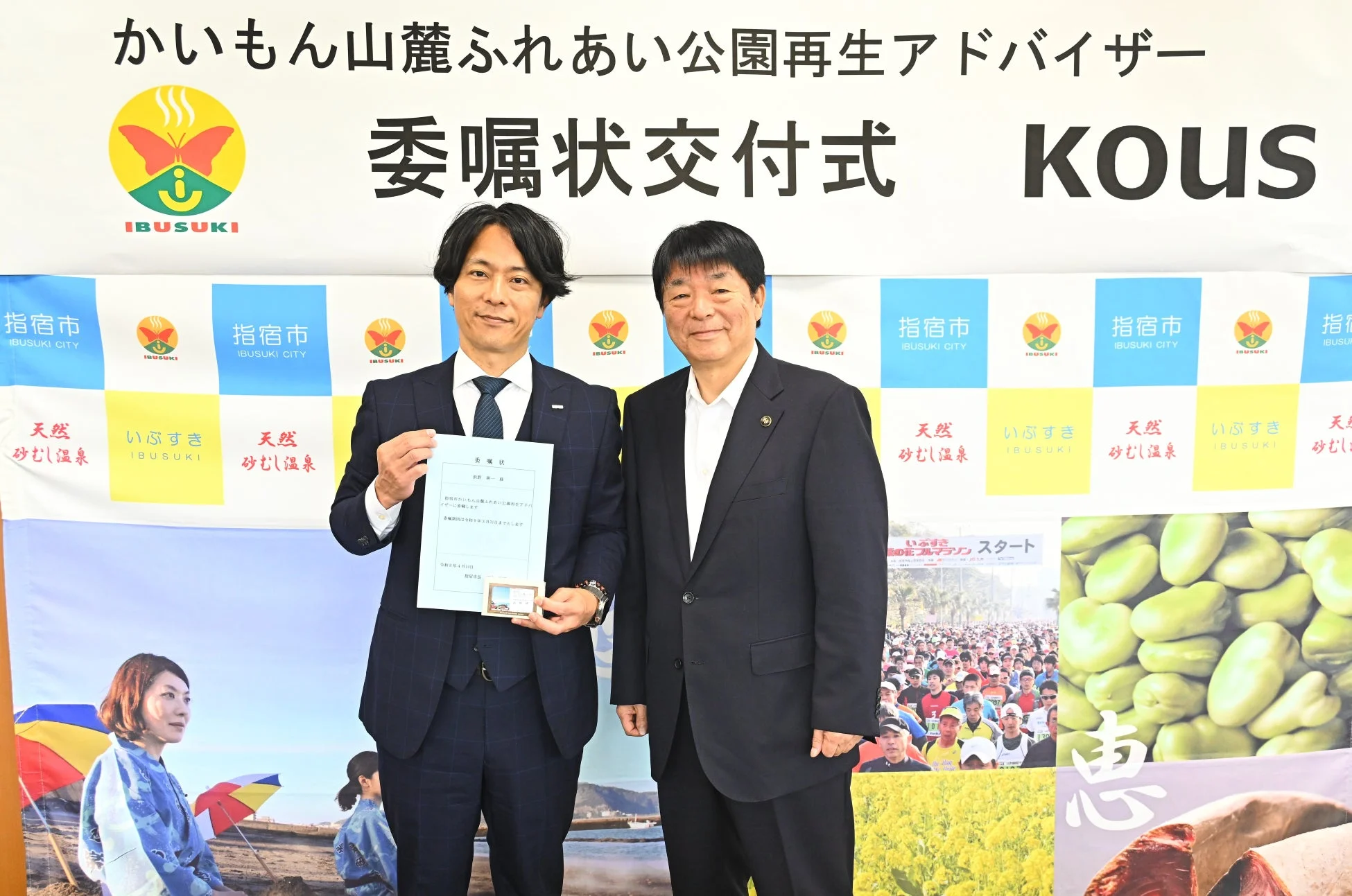 指宿市のかいもん山麓ふれあい公園再生アドバイザー委嘱状交付式で、二人の男性が証書を手に立っている。背景には指宿市の観光をPRする多様なイメージが描かれている。