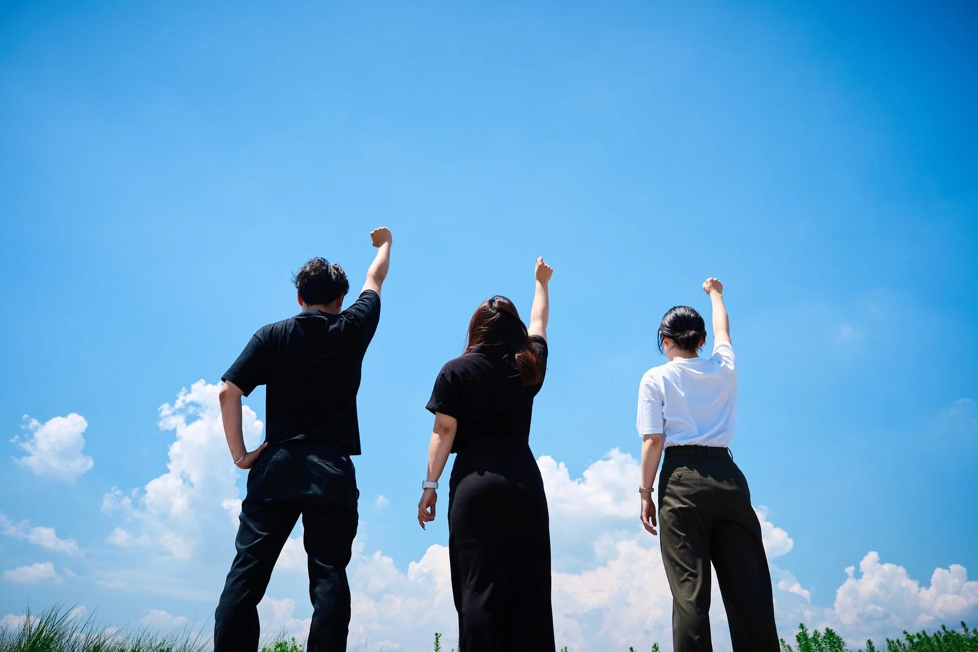 青い空と白い雲を背景に、3人の人物が後ろ姿で拳を高く掲げている様子。