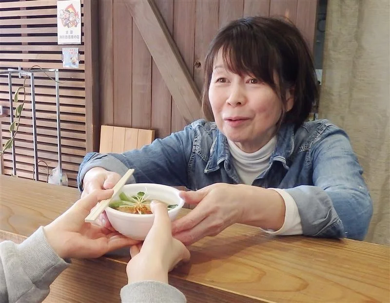 木製カウンターに座る女性が、別の人物から箸が添えられた小さな白い器を受け取っている様子です。