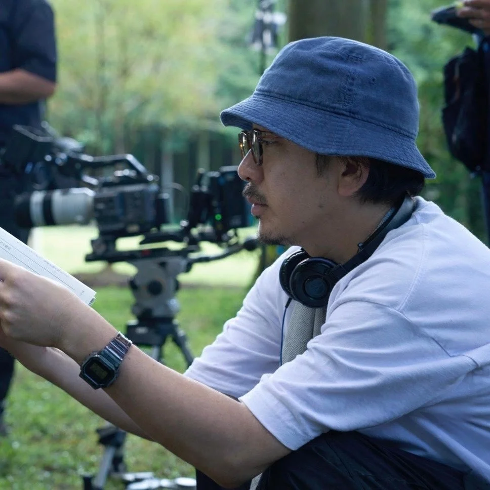 青いバケットハットをかぶった男性が、屋外の撮影現場で台本のような書類を真剣な表情で読んでいる様子。