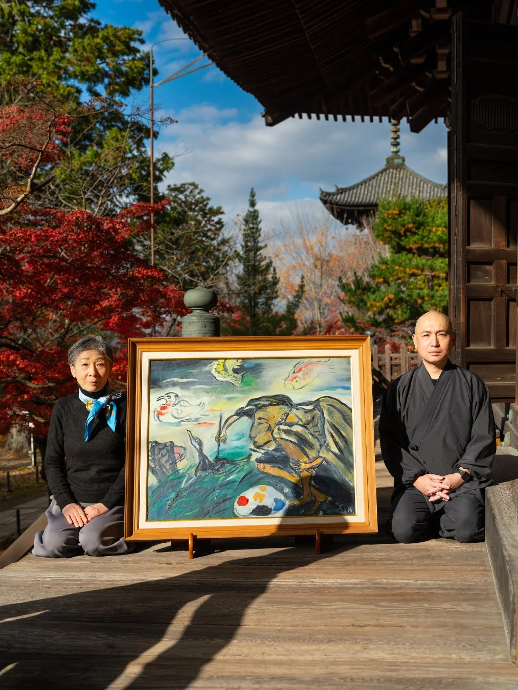 紅葉の寺院で女性と僧侶が絵画を挟んで座る様子