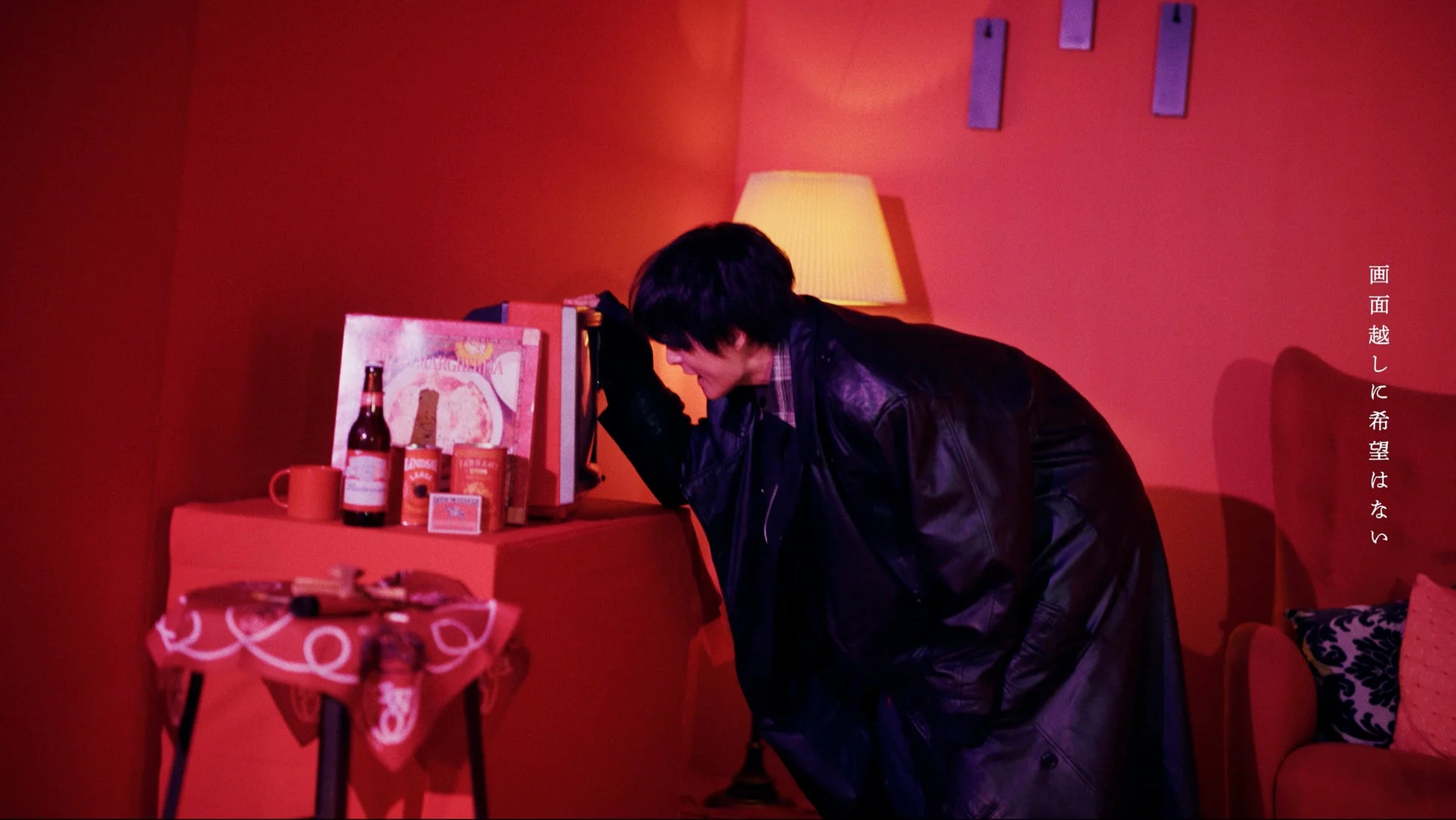 赤い部屋で黒いレザージャケットを着た男性が、テーブル上のビール瓶や缶に身を乗り出している。画面の右側には「画面越しに希望はない」という日本語の文字があり、全体的に暗く、どこか物憂げな雰囲気が漂っている。
