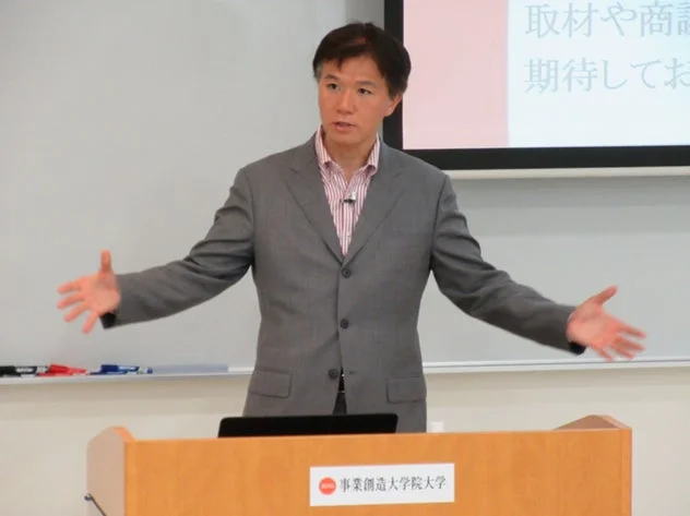 スーツを着た男性が壇上でプレゼンテーションを行っています。背後にはプロジェクタースクリーンがあり、一部テキストが見えます。壇上には「事業創造大学院大学」のロゴがあります。