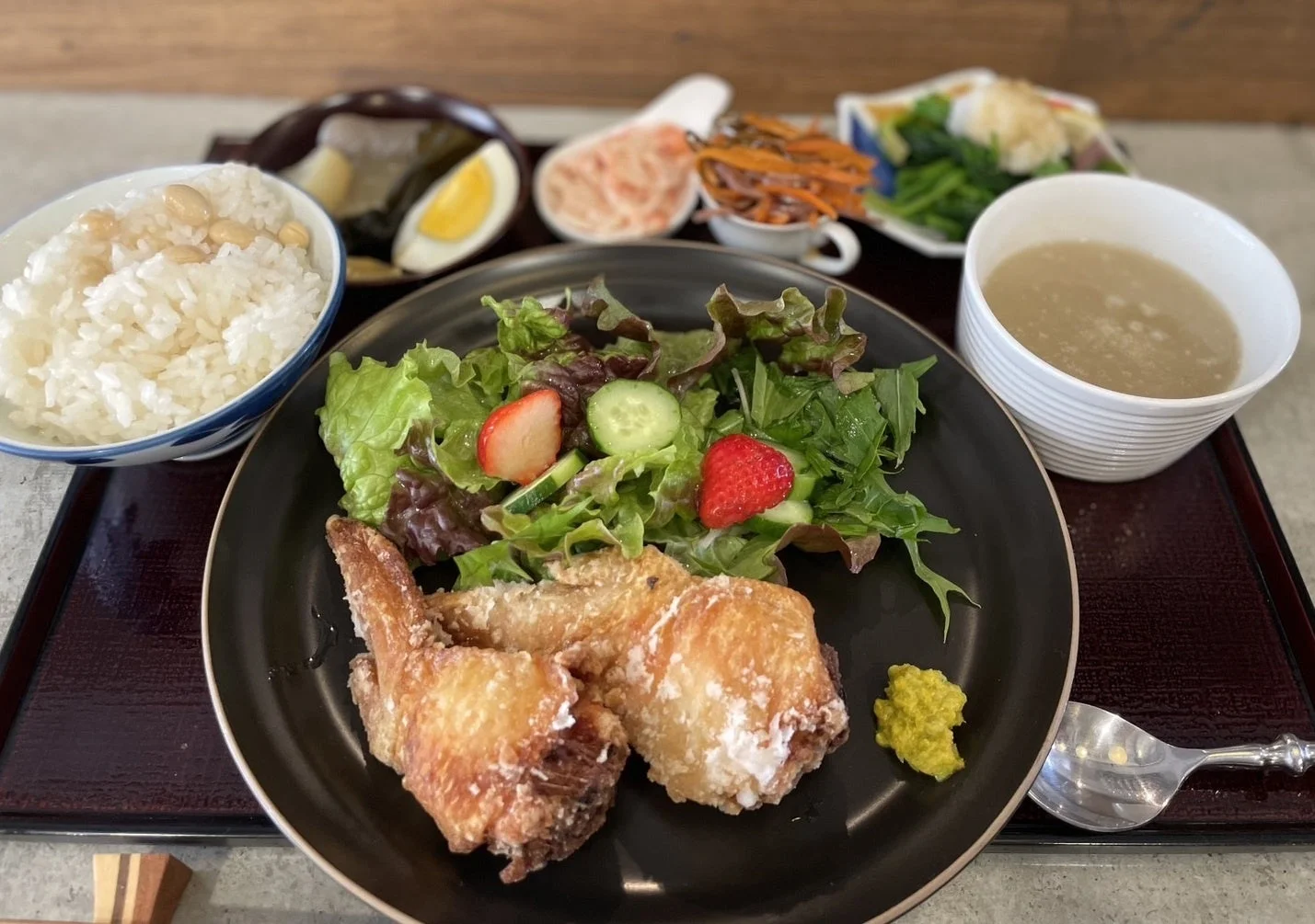 揚げた鶏肉と新鮮なサラダがメインの定食。ご飯、スープ、複数の副菜が添えられ、バランスの取れた和食の食事がトレイに並べられています。
