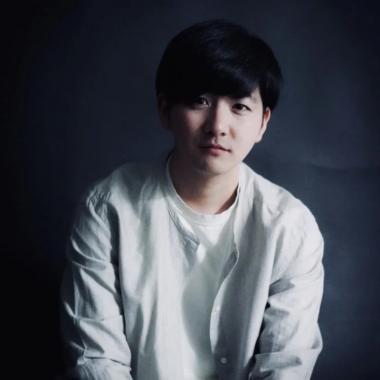 暗い背景の前で、薄い色のスタンドカラーシャツを着た若いアジア人男性のポートレート。