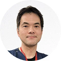 白背景を背に、紺色のスクラブを着用し、赤いストラップを首から下げたアジア人男性の顔のアップです。彼はカメラをまっすぐ見ており、真剣な表情をしています。
