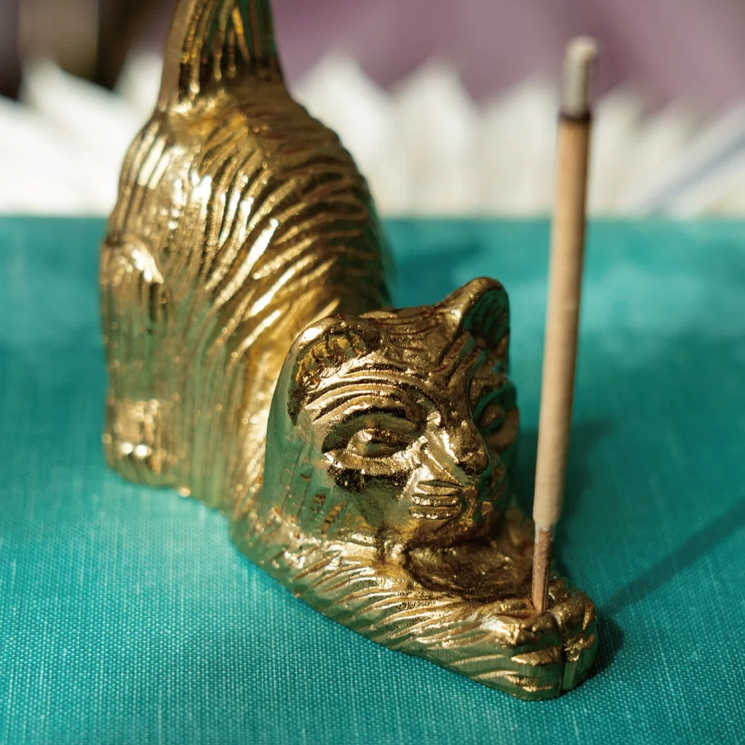 青緑色の布の上に置かれた、金色の猫の形をしたお香立てのクローズアップ。猫の足元にお香が立てられており、落ち着いた雰囲気と装飾的な美しさを感じさせます。