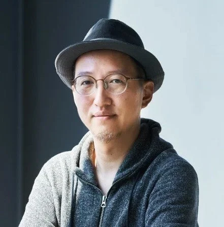 帽子と眼鏡をかけた男性のポートレート。グレーのパーカーを着ており、カメラ目線で穏やかな表情をしている。背景はシンプルで、カジュアルな雰囲気の一枚です。