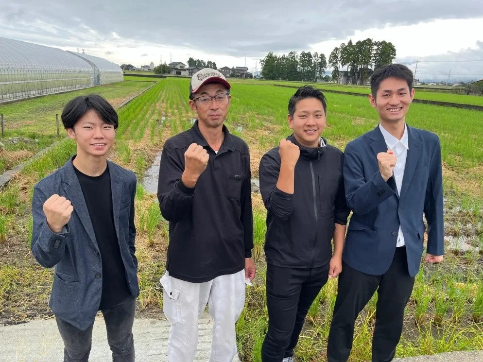 田んぼと温室を背景に、4人の男性が笑顔で拳を握り、意気込みを示すポーズをとっています。農業や地域活動に関わるグループのようです。