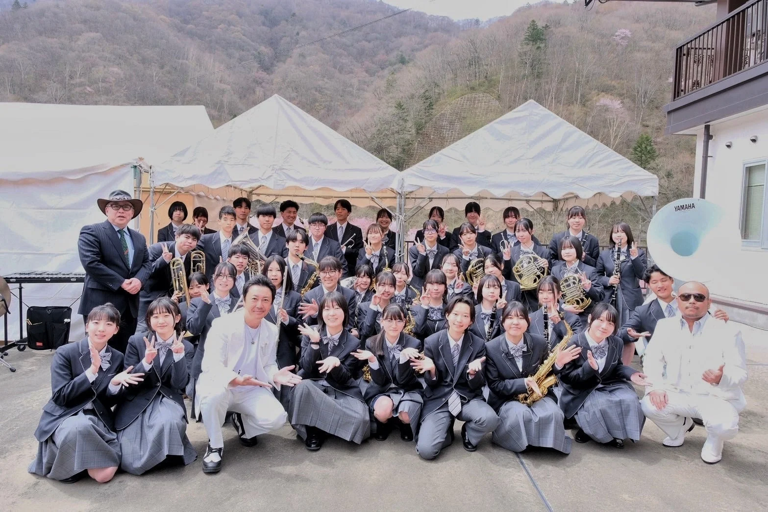 制服姿の学生と数人の大人が、様々な楽器を持って屋外で集合写真を撮っています。吹奏楽部の活動かイベントの一幕と思われ、皆笑顔でポーズをとっています。背景には山と白いテントが見えます。
